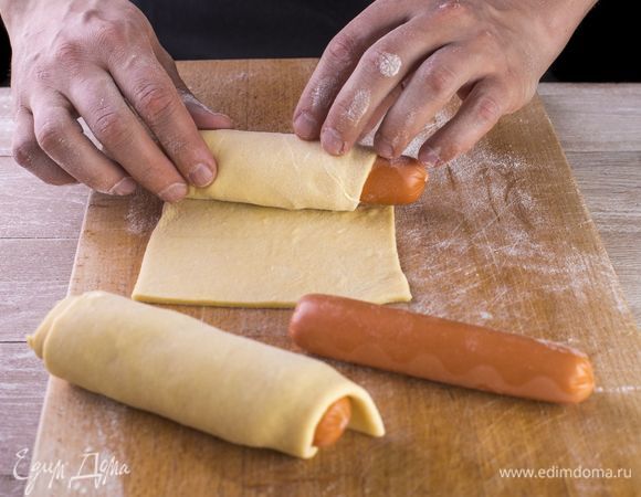 Положите сосиску на пласт теста, отступив от края 2–3 см. Плотно заверните сосиску в «кокон». По такому же принципу соберите остальные сосиски в тесте.