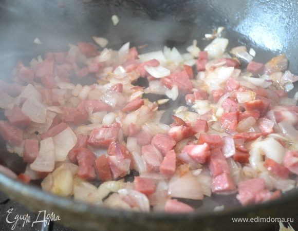Как только лук станет румяниться, добавьте мелко нарезанную копченую колбаску. Жарьте минуту, помешивая. Колбаса отдаст свой аромат копчения. Затем добавьте измельченный чеснок.