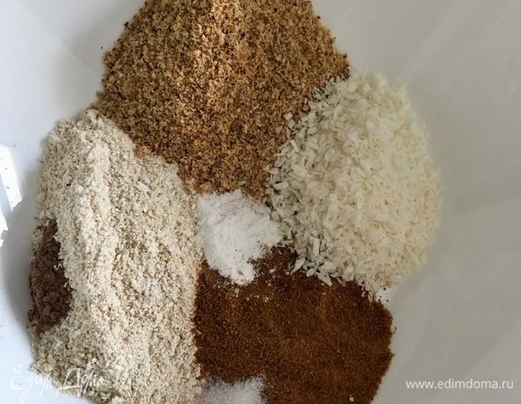 Теперь в большой миске необходимо смешать все сухие ингредиенты. Фигурной россыпью у меня пока не получается, но так тоже вроде видны все сухие составляющие морковной основы. Итак, по порядку. Слева у меня самодельная овсяная мука с чайной ложкой корицы. Сверху — фундучная мука (можно заменить на миндальную или другую ореховую муку). Справа — кокосовая стружка. Снизу насыщенного коричневого цвета — это кокосовый сахар. И по центру чайная ложка разрыхлителя. Итак, смешать все сухие ингредиенты.