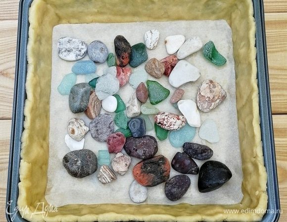 Прошло 30 минут, тесто отдохнуло, и пора им заняться. Но сначала включаем духовку на 200°С, пусть разогревается. Тесто раскатываем в тонкий пласт, выкладываем в квадратную форму (24х24 см), формируем бортики, от души накалываем вилкой, застилаем пергаментной бумагой. Сверху кладем груз. Владивосток — город у моря, поэтому в ход идут все морские дары, я набрала небольшие камешки и прекрасно обточенные стеклышки, тщательно их вымыла и теперь использую в качестве груза. Но горох и фасоль тоже вполне подойдут. Отправляем в уже разогретую духовку наше тесто на 10 минут.