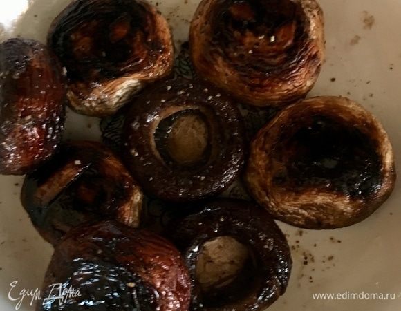 Начнем приготовление салата с грибов. И не просто грибов, а шампиньонов гриль. Это отдельная песня данного салата. На фото вот эти черненькие немного обугленные шляпки — уже готовые шампиньоны гриль. Я не успела сделать пошаговых фото их приготовления, к сожалению. На решетке их готовил мой папа, пока мы с мамой занимались огородными работами, придется поверить мне на слово. Берем крупные шляпки шампиньонов, моем, обсушиваем бумажным полотенцем. Удаляем аккуратно ножку, не повредив шляпки. В каждую шляпку кладем маленький кусочек сливочного маслица и присаливаем немного. Теперь ставим грибочки на решетку и жарим до готовности. Масло начинает кипеть в шляпе, пропитывая грибок своими соками. Снимаем готовые шляпки также отверстиями вверх. Вообще, такие грибы едят как самостоятельную закуску следующим образом. Сначала выпивают сок из шляпки и только потом закусывают самим грибком. Вкусно до жути! Несколько таких шляпок предлагаю оставить целенькими для украшения салата, остальные нарежем.