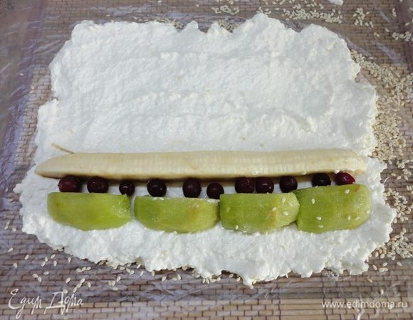 Коврик застелите пленкой и посыпьте кунжутом или кокосовой стружкой (у нас, в семье любителей, таковой не нашлось), выложите творожную массу с помощью мокрой ложки, затем по краю расположите фрукты, а также какую-нибудь красненькую ягоду. У меня брусника. Можно гранат, смородину и т. п.