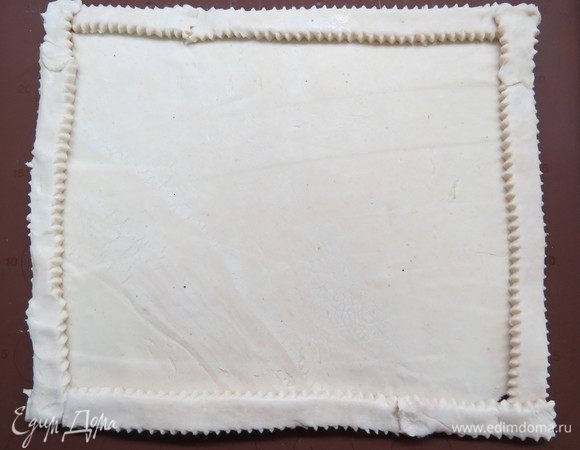 Пласт слоеного теста раскатать на силиконовом коврике или бумаге для выпечки, по периметру сделать бортики.