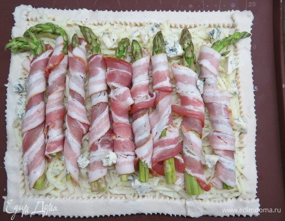 У спаржи отрезать нижнюю твердую часть, обмотать каждый стебель кусочком бекона, равномерно выложить поверх сыра.