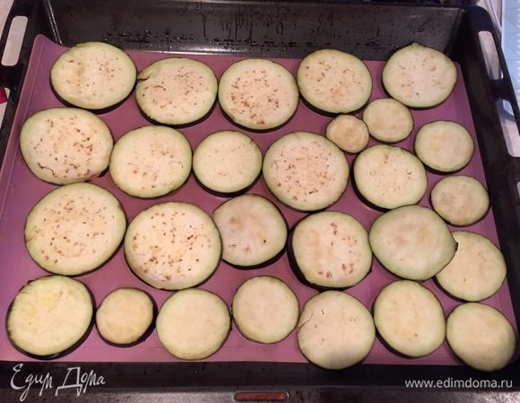 Выложить кружочки баклажанов на противень, застеленный пергаментом. Сверху на каждый баклажан выложить несколько кусочков чеснока.
