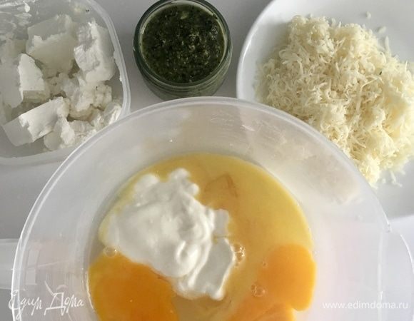 Приготовим заливку пирога, для которой мы как раз и готовили ароматный соус песто. Для этого в миске смешайте три яйца, три столовых ложки натурального йогурта. Перемешайте вилочкой. Натрите моцареллу для пиццы на мелкой терке. И подготовьте фету.