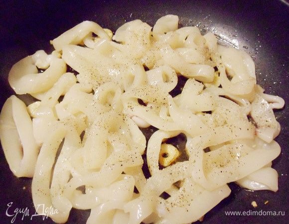 На ту же сковороду с вытопившимся жиром от бекона выложить нарезанный пластинками чеснок. Увеличить огонь до среднего уровня и обжаривать чеснок, помешивая, около 30 секунд. Затем выложить на сковороду кольца кальмаров.