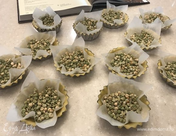 Из бумаги для выпечки вырезать квадраты по количеству и размеру тарталеток, выложить бумагу на тесто и насыпать сверху горох. Выпекать тарталетки с горохом в заранее разогретой до 190°C духовке 5–7 минут.