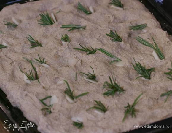 Противень смазать 1 ст. ложкой оливкового масла, равномерно выложить тесто, сделать в нем небольшие углубления и воткнуть в них чеснок и розмарин.