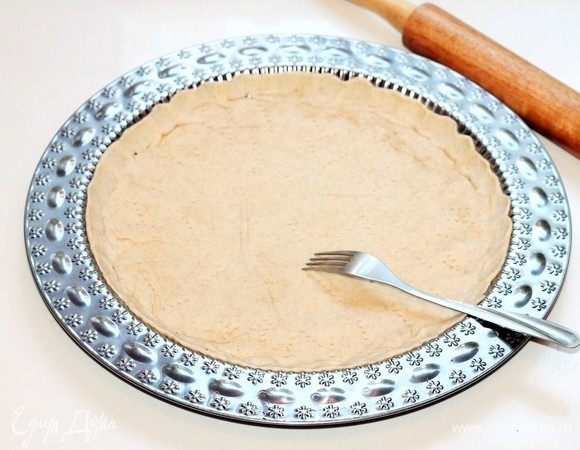 Обмять и тонко раскатать тесто на припыленной мукой столешнице в круг диаметром 26 см, и при помощи скалки перенести в смазанную маслом формочку (диаметр = 26 см). Проткнуть тесто вилкой в нескольких местах.