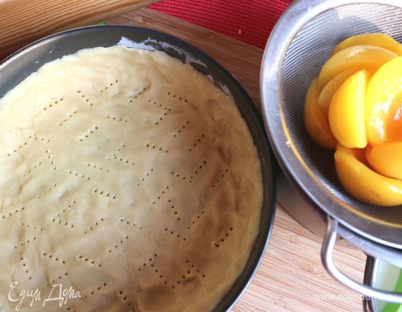 Раскатать тесто скалкой на присыпанной мукой поверхности. Часто это делают между двумя слоями пергаментной бумаги, потом бумага убирается. Разъемную форму смазать небольшим количеством масла (излишки удалить салфеткой), обсыпать мукой, лишнюю муку стряхнуть. Поместить тесто в форму, сделать невысокие бортики из теста. Основу из теста наколоть вилкой, убрать в холодильник минимально на 20 минут, лучше — больше. Консервированные персики откинуть на сито. Строп не понадобится.