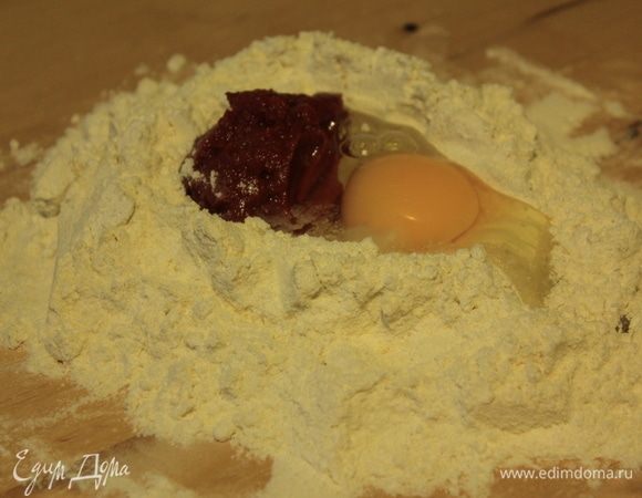 Готовим красное тесто для пасты. Смешиваем оба вида муки, высыпаем на рабочую поверхность и делаем углубление. Разбиваем туда яйцо, добавляем томатную пасту и начинаем замешивать тесто. Сначала вилкой, а потом руками.