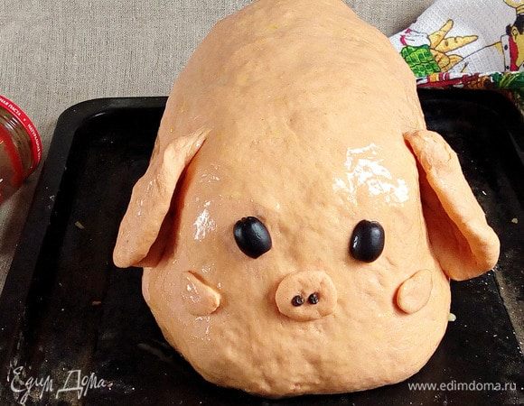 От оставшегося теста отщипнуть кусочек размером с два грецких ореха. Из него сделать ушки, щечки, пятачок и хвостик. Оставшийся объем теста раскатать в овальный сочень, которым накрыть всю галетную башенку. Края основы поднять кверху по периметру всей башенки, а обтяжку подвернуть под дно основы. Прикрепить ушки, пятачок, щечки, глазки (оливки) и ноздри (перец черный горошком).