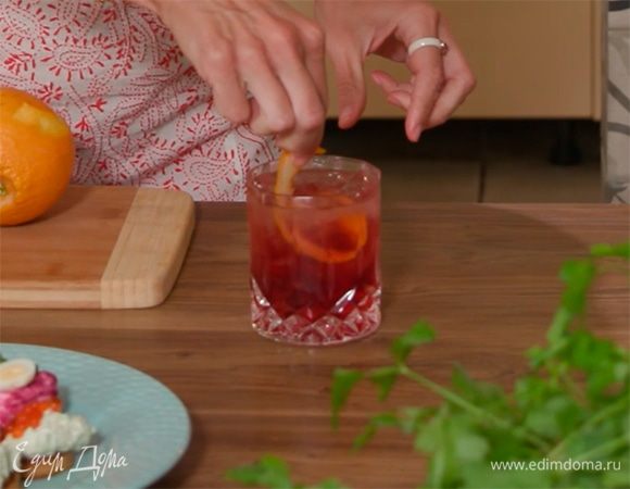 С апельсина двумя тонкими полосками срезать цедру, одну из них поместить в бокал с коктейлем, другую выжать в напиток.