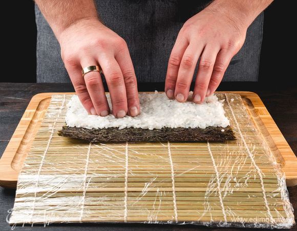 Лист нори выложите на циновку, накрытую пищевой пленкой. Смочите руки холодной водой и распределите рис на нори ровным слоем.