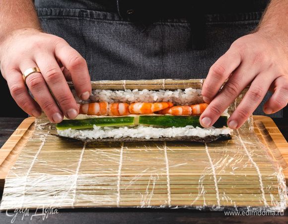 Сверху в ровную линию выложите креветки, мягкий сыр и нарезанный соломкой огурец. Скрутите ролл при помощи циновки, удалите пищевую пленку, разрежьте на несколько частей.