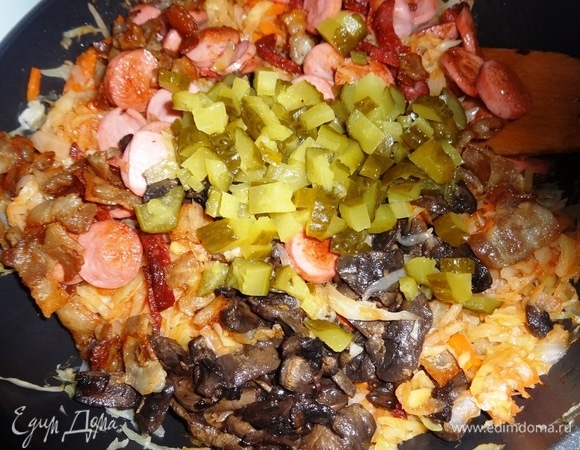 В сковороду с капустой добавить соленые огурцы, обжаренные сосиски, колбасу, бекон, грибы вместе с жиром, перемешать.