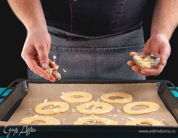 Противень застелите пекарской бумагой, выложите тесто в кондитерский шприц и сформируйте основу для пирожных, выдавливая тесто в форме колец. Сверху посыпьте заготовки по кругу миндальными лепестками. Поместите противень с пирожными в разогретую духовку и, понизив температуру до 180°С, выпекайте 15 минут, затем уменьшите температуру до 170°С и выпекайте еще 15 минут. Затем выключите духовку и оставьте пирожные еще на 5–7 минут в духовке.