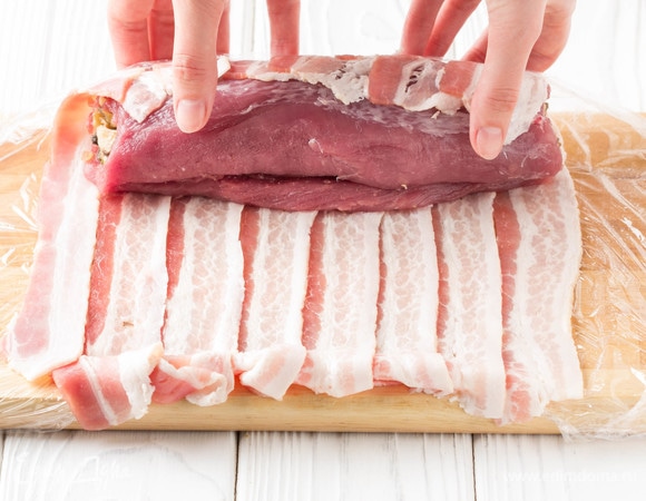 На доску положить пищевую пленку и внахлест выложить ломтики бекона. На один край положить свиной рулет и с помощью пленки завернуть его в бекон.