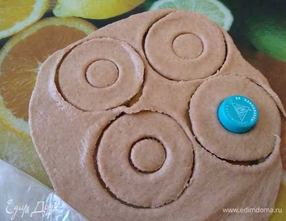 Тесто раскатать в пласт толщиной 0,5 см и широким стаканом вырезать кружки. Выемкой меньшего размера (у меня крышка от бутылки) из каждого кружка вынуть середину, придавая таким образом тесту форму пончика.