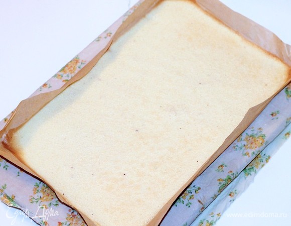 Выпекаем примерно 15 минут, не пересушиваем. Ориентируйтесь на свою духовку. У меня небольшие противни размером 22x32 см, и я выпекала 2 тонких коржа. На стол кладем полотенце, сверху на него — готовый горячий бисквит с пекарской бумагой. Острым ножом обрезаем края рулета с двух сторон, чтобы при скручивании не ломался.