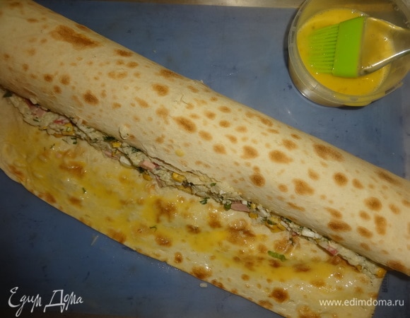 Выложить лаваш на противень с силиконовым ковриком. Свернуть лаваш с начинкой в рулет (не до конца). Яйцо взболтать. Смазать внутренний край и остальную поверхность яйцом. Боковые части немного завернуть внутрь.