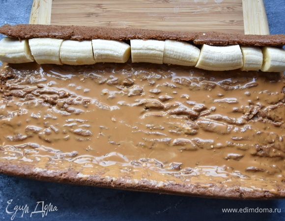 Рулет раскрутить. Освободить его от пергаментной бумаги, убрать кухонное полотенце. Пропитать бисквит любым сахарным сиропом. Выложить большую часть крема. На край выложить очищенные от кожуры бананы.