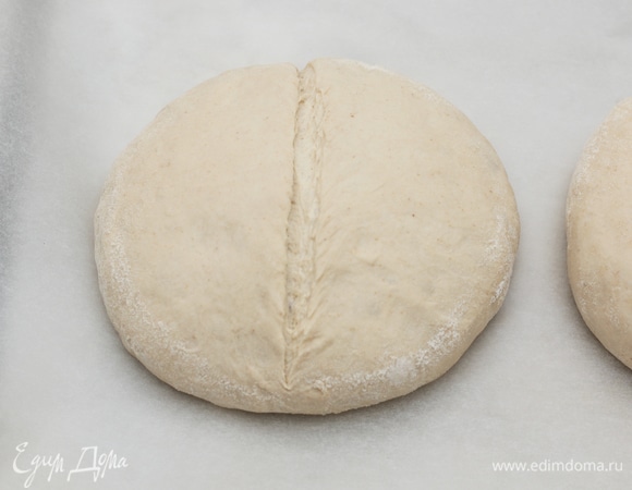 За полчаса до окончания расстойки включите духовку на 230°C, чтобы она как следует прогрелась. На дно духовки установите старую пустую сковородку, чтобы она прогрелась вместе с духовкой. После расстойки тесто увеличится в объеме и станет более мягким. Переложите его швом вниз на противень, застеленный бумагой для выпечки. Сделайте надрезы. Надрезы помогают тесту лучше и правильнее пропечься, поэтому надо наловчиться их правильно делать, я пока только учусь. Но могу сказать, что это лучше всего делать обычным бритвенным лезвием, держа его под углом примерно в 30°C к тесту. Глубина надреза, около 5–7 мм.