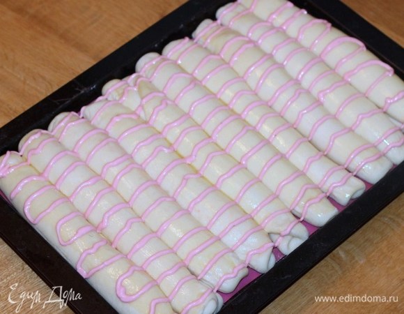 Достаем противень с заготовками и переключаем духовку на 180°C. Смазываем булочки взбитым желтком. Сверху рисуем на заготовках полоски малиновым кремом.