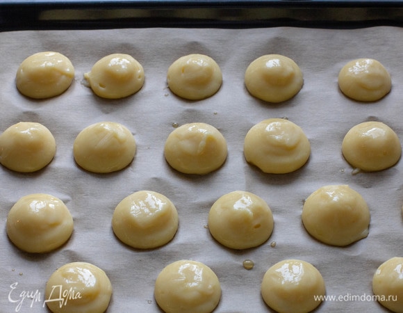 Тесто отсадить из кондитерского мешка на противень, застеленный бумагой для выпечки. Можно использовать любой прочный пакет, срезав уголок, чтобы отверстие в диаметре было примерно 1,5 см. При необходимости влажными пальцами сгладить хвостики. Взбить яйцо со щепоткой соли и смазать им сверху профитроли.