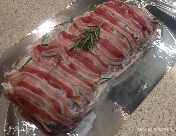 Шпигуем мясо морковью, чесноком, смесью из соли. Хорошо натираем сметаной с солью. Накрываем полосками бекона. Посыпаемся розмарином. Ставим на один час в холодильник.