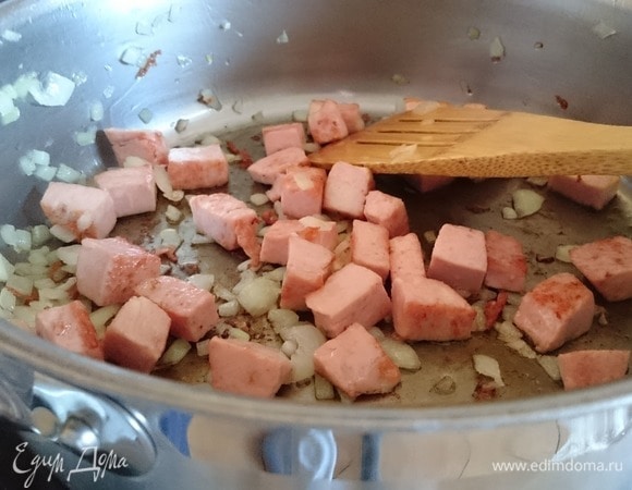 Нагрейте масло в большой сковороде. Положите нарезанную колбасу и жарьте на среднем огне. К чуть обжаренной колбасе добавьте лук и готовьте до золотистого цвета около 3 минут.