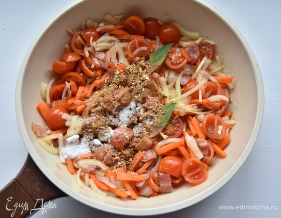 К бекону добавить измельченный чеснок, нарезанные соломкой лук, морковь и томаты. Посолить, добавить специи по вкусу. Обжарить овощи минуты 3–4.