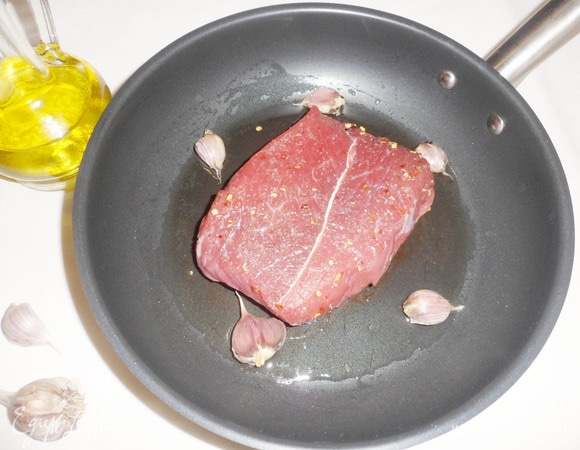 На сковороду налить оливковое масло, добавить раздавленные зубчики чеснока. Разогреть масло и выложить мясо.