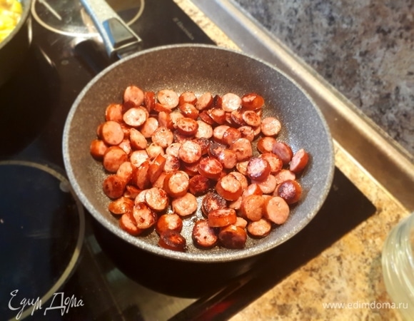 На отдельной сковороде обжарить копченые колбаски. Сначала на медленном огне, чтобы вытопился жир, затем огонь увеличить для получения румяной корочки.