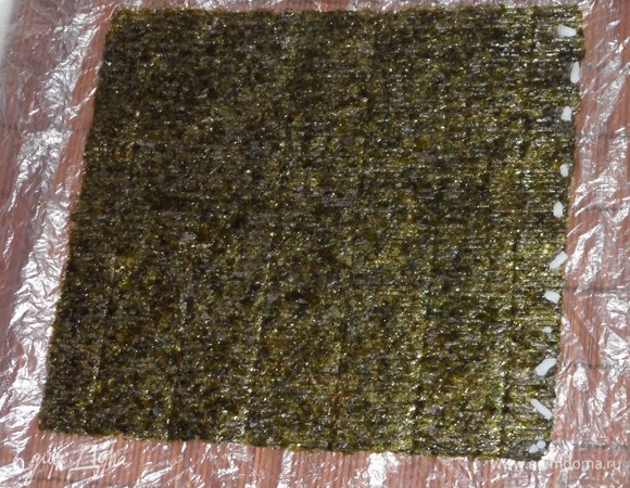 Циновку застелить целлофаном. Положить на него лист нори. С одного края положить несколько рисинок. Они нужны для крепкого скрепления.