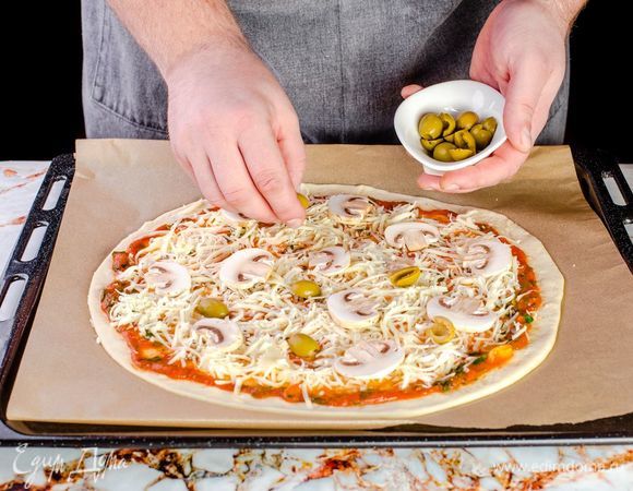 Сверху на пиццу выложите натертый сыр, нарезанные шампиньоны и разрезанные пополам оливки.