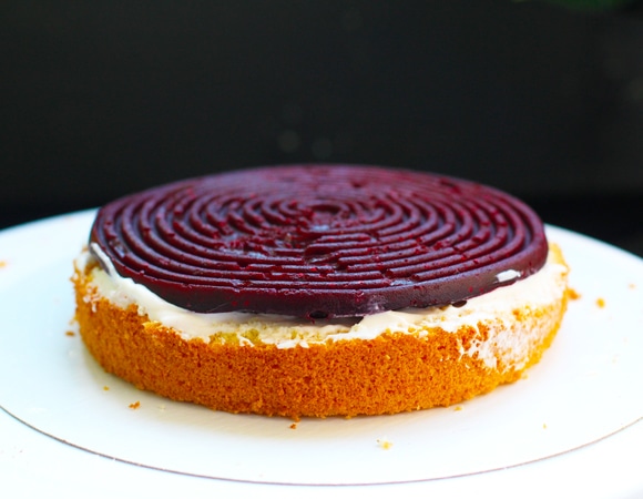 Сборка. На подложку намазать ложку крема (чтобы торт не ездил по коробке), сверху положить первый бисквит, пропитать, если считаете нужным, слегка смазать кремом. Сверху уложить первый кружок конфи. Далее поставить вокруг коржа разъемное кольцо и ацетатную пленку и собирать уже в нем, так как йогуртный крем очень нежный и форму не держит. Выдавить из мешка равномерно треть йогуртового крема, далее — бисквит, пропитка, еще треть крема. Уложить снова корж, смазать чуть кремом, затем — кружок конфи и оставшийся крем.