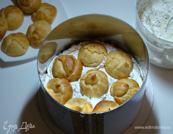 На сервировочное блюдо установить одну часть двойных коржей. Сверху аккуратно установить кольцо, внутри проложить ацетатной или пищевой пленкой. На коржи выложить половину сливочного крема и уложить охлажденные профитроли, слегка вдавливая их. Сверху равномерным слоем выложить оставшуюся часть крема.