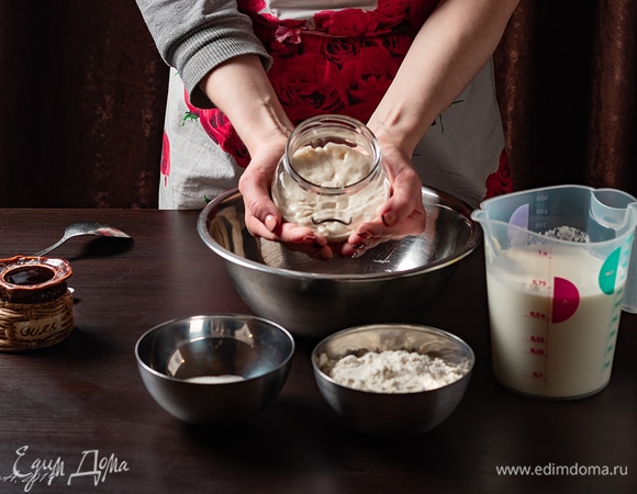 Для блинов нам потребуются пшеничная закваска, мука, теплое молоко, сахар и соль. Сначала мы вырастим пшеничную закваску. Это довольно несложно, просто требует времени и соблюдения некоторых условий. Нам потребуется 25 г муки пшеничной и 25 г воды. Воду берите либо отстоявшуюся, либо фильтрованную, на крайний случай — холодную из-под крана, но ни в коем случае не кипяченую. Скорость роста закваски не зависит от количества ингредиентов, я просто делала «на пробу», а вы можете сделать побольше, тогда просто возьмите столько, сколько хотите, главное, чтобы в одинаковом количестве. Смешайте так, чтобы не было комочков, положите в стеклянную банку и плотно закройте крышкой. Капроновой или винтовой — без разницы. Будущую закваску отправьте в теплое место приблизительно на сутки. Температура около 28°C. Сразу оговорюсь: вариантов может быть несколько, я делала так, как меня учила знакомая, у которой большой опыт выпечки на закваске. Поэтому ознакомиться с какими-то нюансами, почитать дополнительный материал можно и нужно, потому что это затягивает, раз попробуешь, и понеслось… Примерно через сутки масса пойдет пузырями, разжижится, увеличится в объеме и приобретет специфический запах. Не следует пугаться, все идет по плану. У нас начались процессы брожения. Теперь нам надо ее покормить. Содержимое банки слегка перемешать, отделить 25 г (я пользуюсь весами). Остаток залить кипятком, слить, тщательно вымыть банку (без использования средств, просто водой), высушить. В закваску долить 25 г воды и положить 25 г муки пшеничной. Тщательно размешать. Тесто должно быть нежидким и в идеале тянуться ниточками при размешивании. Не замешивайте очень круто — наши бактерии еще слабенькие и могут не справиться с работой и задохнуться. Также не надо разводить жидко, иначе бактериям в стадии активного роста будет нечего есть, и закваска не будет расти. Так что количество муки может быть и чуть больше, и чуть меньше, но 1:1 — это классика. Снова поставьте в теплое место, пока до утра. Поздравляю, у вас теперь живет маленькое существо, которое требует заботы и любви. Процесс созревания закваски — вещь индивидуальная. При совпадении всех факторов уже наутро после первого кормления будет видна работа бактерий — закваска по-прежнему пускает пузыри, чуть увеличивается в объеме, а, может, и не чуть, поэтому не ставьте ее там, где очень тепло, все-таки бактерии — это бактерии, в закваске есть и полезные, и вредные, которые под воздействием тепла могут одержать победу. Если процессы активные, покормите ее по той же схеме. Если видимых изменений не произошло, оставьте до вечера, если по-прежнему все тихо, все равно кормите. А на следующий день режим питания должен быть двухразовым: утро/вечер, с интервалом примерно 12 часов. У моей наставницы чисто пшеничная закваска вызревает до готовности за 4 суток. Мне же потребовалось больше недели. И то, и другое — норма. Возможное устранение проблем с закваской: «Что делать, если она не растет и не проявляет видимой активности?». Запомните: если есть хоть немного пузырьков, кислый запах, похожий на кефир, значит ваша закваска жива, просто ее надо завести. Во-первых, продолжайте кормить, но смените муку и воду. Лучше сразу взять цельнозерновую муку или первого-второго сорта, а если такую не нашли, берите высшего сорта с количеством белка от 11 (просто посмотрите состав). Воду пробуйте разную, за исключением газированной, кипяченой и сильно очищенной. Мне подошла вода из скважины. Во-вторых, проверьте помещение, где стоит банка на предмет сквозняков и перепадов температуры (может быть, кухня за ночь остывает, и закваска мерзнет). В-третьих, попробуйте замесить ее гуще. Возможно, бактериям просто не хватает питания, они съедают то, что им дали, и засыпают. Пропорции кормления в этом случае следующие: на 25 г закваски — 20 г воды и муки такое количество, чтобы тесто получилось по консистенции похожим на модный нынче сквиш (такая игрушка, которую очень приятно мять). Вы можете бережно катать этот комочек между ладоней, он не липнет и хорошо держит форму. Все вышеперечисленные хитрости применены лично мной. В процессе созревания закваска может пахнуть сильно, в интернете сравнения самые разные. Верными признаками готовности закваски станут кремовый цвет, много пузырьков, кислый вкус, стабильный рост после кормления (в идеале вообще 4–6 часов) и приятный запах. Как его описать, я не знаю, похож на яблочное пюре. Но! Если ваша закваска покрылась плесенью, стала пахнуть затхлостью (именно запах «старья»), изменила цвет на зеленоватый или синюшный — это значит, что, увы, она погибла, и придется начинать заново. Такое случается, если изначально была плохая зараженная мука или вода (поэтому осторожнее с водой из колодцев и скважин, тем более сейчас, когда весна), или при грубом нарушении технологии (например, забыли в теплом месте и при этом пропустили несколько кормлений).