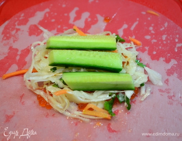 На смоченной в воде рисовой бумаге распределить икру, далее — горсть квашеной капусты с зеленью, дольки огурцов.