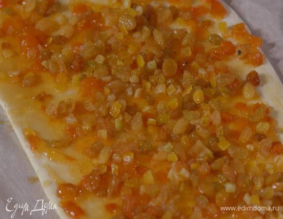 Предварительно размороженное тесто слегка раскатать, равномерно смазать вареньем и посыпать изюмом с цукатами, а затем прижать начинку руками.