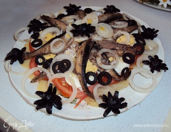 На плоское блюдо выложить слоями картофель, помидоры, шпроты, яйца. Все слои, кроме рыбы, немного посолить и поперчить. Полить салат масляно-уксусной заправкой. Украсить колечками лука, оливок и фигурками из оливок.