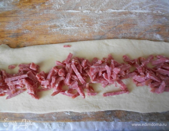 Из полученного теста оторвать кусочек весом примерно 100 г. Остальное тесто разделить на 3 одинаковые части. Каждую часть теста раскатать в прямоугольник примерно 70–80 см длиной и 8–9 см шириной. Выложить на них начинки: на первую — колбасу.
