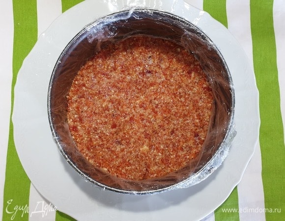 Форму для выпечки обернем пищевой пленкой. Утрамбуем основу. Убираем в холодильник, пока занимаемся основным слоем.