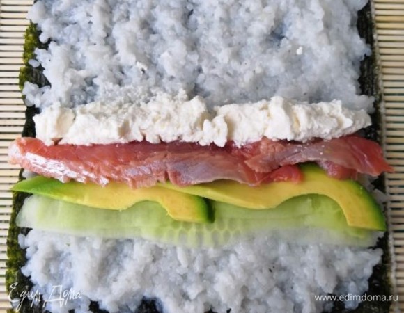 На коврик выложить лист нори пупырышками вверх. На нем, смочив руки водой, распределить рис. Далее выкладываем начинку. В первом случае это огурец, авокадо, красная рыба и творожный сыр.