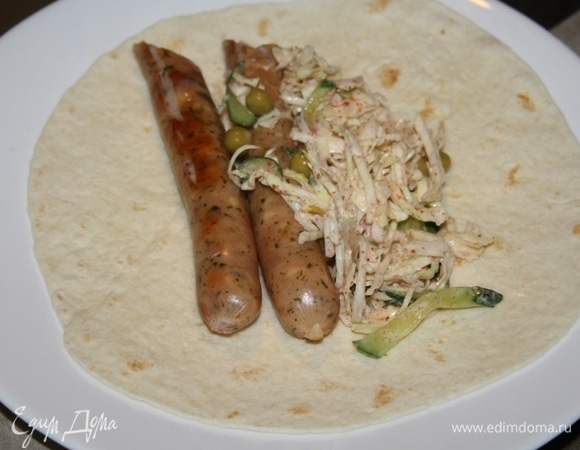 Колбаски разрезать пополам, выложить на тортильи, добавив капустный салат.