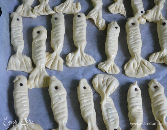 Соедините края, как у пирожка, придайте форму рыбки. Зубочисткой сделайте надрезы в виде чешуи, а из кусочков маслин (черный перец) — глазки. Уложите рыбок швом вниз на противень, покрытый пекарской бумагой. Смажьте яйцом и выпекайте в духовке при 180°C до золотистого цвета.