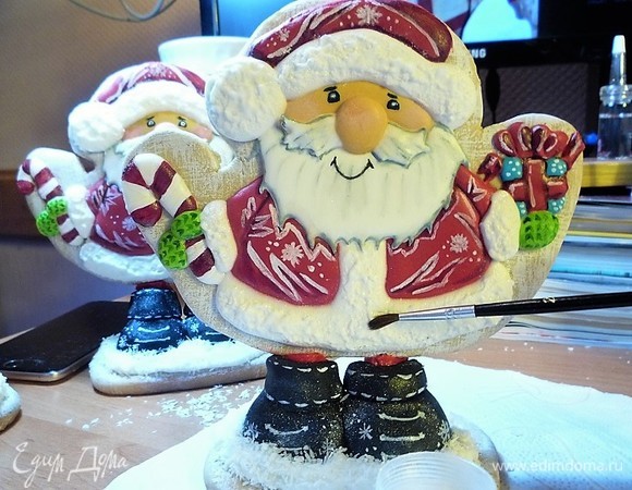 Полностью высохший пряник прорисовываем с помощью тонкой кисти, белой краской разрисовываем шубку, делаем точечки на коробке, прорисовываем красной краской конфету в руке, черную краску мешаем с белой и прорисовываем усы и бороду, зеленой краской раскрашиваем варежки, рисуем глазки и ротик, сухой розовой краской делаем румянец на носу и щеках. Делаем имитацию пушистой шапки и шубы. Для этого в палетку из кондитерского мешка выкладываем немного айсинга, окунаем в него кисть и макающими движениями наносим айсинг на низ шапочки, бубон и середину и низ шубки.