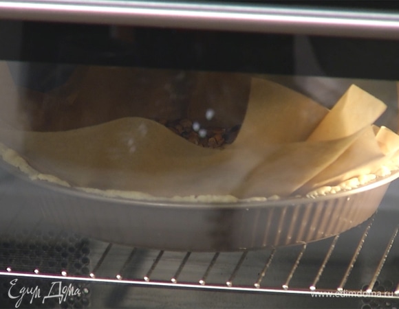 На тесто выложить пергамент, сверху заполнить форму бобами — используйте 1–1,5 кг. Корж выпекать при 180°С в течение 20 минут, почти до готовности.